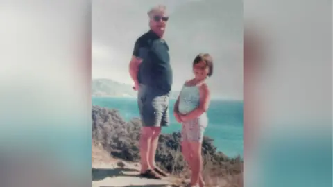 Suzanne Wiggins A man stands on a coastal cliff with a young girl. The girl is smiling and wears a colourful outfit. The man wears sunglasses and has grey hair. Looks to be abroad due to blue sea in background and both wear shorts