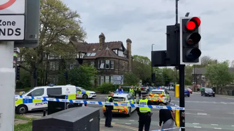 Cathy Killick/BBC A police cordon in place at a road junction. Several uniformed officers can be seen behind tape, along with several police vehicles. A traffic light, on red, is in the foreground. 