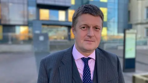 Richard Clewer, pictured wearing a dark three-piece suit, pink shirt and tie. 