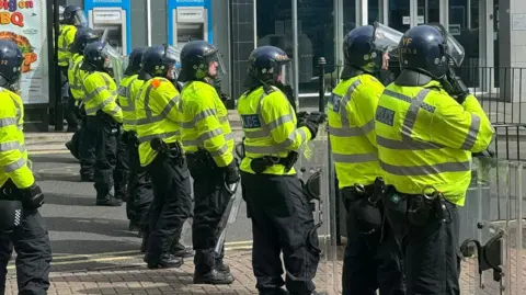 A line of police in riot gear, wearing helmets, fluorescent jackets and carrying riot shields, in a line on a street outside a bank. Ten officers are visible.