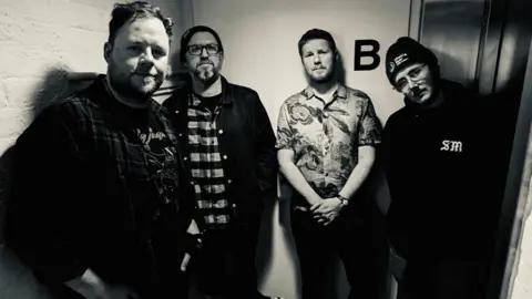 A black and white picture of the four members of Sinews in a corridor, posing for a band photo.