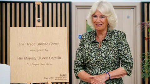 PA media queen Camilla stands smiling next to a memorial plaque on an easel