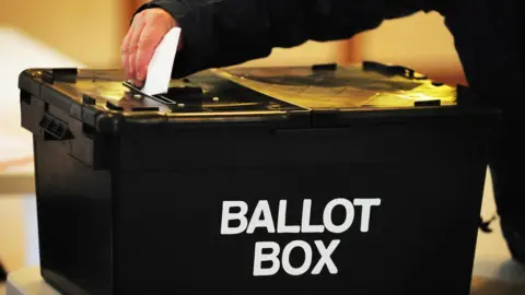 PA Media A large black box that reads Ballot Box, someone is putting a folded up piece of paper in it.