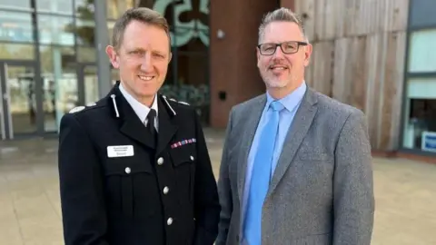 West Mercia Police and Crime Commissioner A man on the left with short brown hair is wearing a black, button up jacket with a silver name badge and stripes of colour on the top of a breast pocket. A man stood next to him on the right has black-rimmed glasses, short grey hair and facial hair, and is wearing a blue shirt and tie and grey suit jacket. 