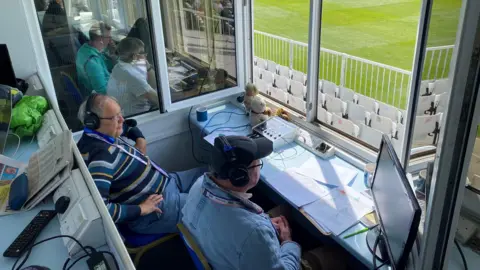 PAUL NEWTON Paul is in a commentary booth with a colleague. They are both looking out to a cricket green, with a monitor on their desk. The photo is taken from above.