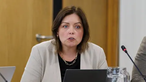 PA Media Deidre Hargey has brown hair down to her shoulders parted in the middle. She is speaking during a committee meeting with a microphone in front of her