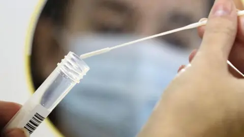 A close-up of the hands of a person placing a cotton swab into a clear test tube in front of a small mirror. In the reflection, you can see the blurred face of the same person wearing a blue mask.