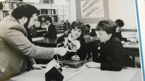 Ackworth School A black-and-white photograph showing an indoor school laboratory in 1986.
Several pupils are seated at tables working with instruments and materials.
In the foreground, a large table occupies the lower half of the image. On the table are multiple pieces of equipment connected by wires. A teacher, a dark haired man with a beard, is pointing to the equipment as pupils look on.
