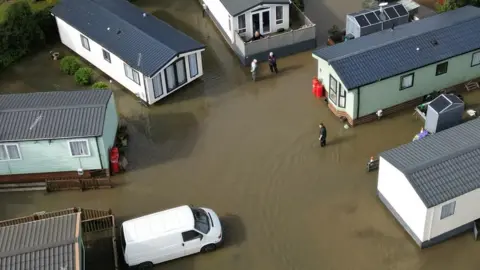 PA Media Mobile homes green and white in colour, with grey roofs, surrounded by water, pictured from the air. A few people are visible standing in the water and there is a white van parked to the bottom left of the picture.