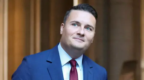 Reuters Wes streeting head shot. He has a blue jacket on, white shirt and red tie. He looks off to the left of the image and has blue eyes and short black hair. 