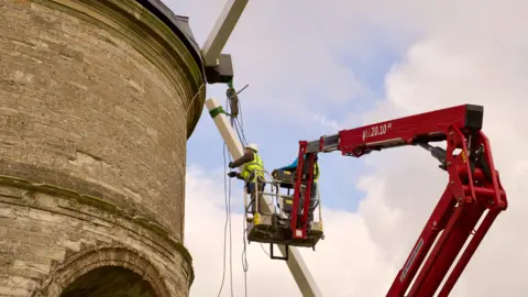 Two men in a red hoist attaching the white stocks to the round stone windmill