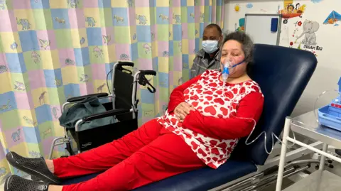 Nikki Fox/BBC A woman in a treatment bay in an urgent treatment centre. She has greying dark hair and is wearing a nebuliser mask, and a red and white top and red trousers. She is sitting on a cushioned blue treatment bed, sitting up with her feet up. Beyond her is a wheelchair and a man in a grey top and wearing a surgical mask. Beyond him is a curtain. 