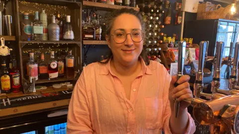 A smiling woman wearing glasses stands behind a bar with one hand on a beer tap with various bottles on the shelves behind her.