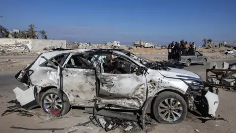 EPA Car wrecks seen following an Israeli strike in southern Gaza. The car is white but slightly burnt. Its roof is missing, it has no windows, and its tires are flat. 