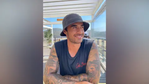 Kelsey Mulcahy An image of a heavily tattooed man wearing a black vest and black bucket hat smiling whilst sitting inside a restaurant on the beach.