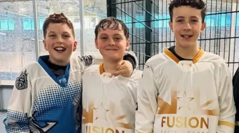 Three boys, Leo, Harry and Archie, standing next to each other at a rink. The one on the far left, wearing a blue and white jersey, has light brown hair. The other two are both in matching gold and white jerseys and have dark hair. All three are smiling.