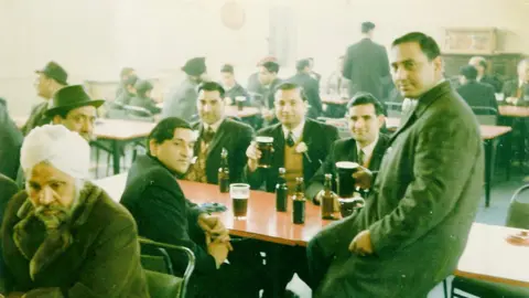 Jagwant Johal Archive photo of Avtar Singh Jouhl on the right with a group of five other men clustered around a table with pints and bottles of beer. It looks like a working men's club in the 1960s