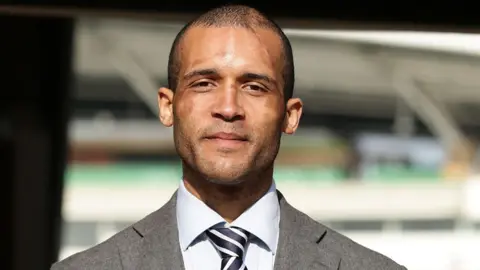 Head and shoulders image of Clarke Carlisle smiling with a sports ground blurred in the background. He is wearing a grey suit jacket, pale grey ad shirt navy and white striped shirt. He has very short dark hair and stubble with scars on his face and head