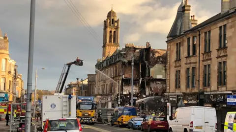 PA Media Fire-damaged tenement building. Firefighters are shooting water into a gap in the building. There is a large emergency service presence. 