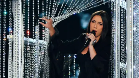 Reuters Yuval Raphael sings on stage, wearing a black dress and jacket. She holds open a glass beaded curtain.  
