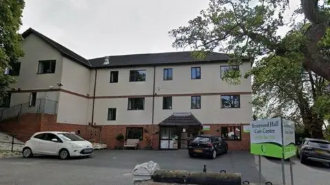 Image of the outside of a care home building, three floors high, mostly painted cream. Three cars are parked on the car park outside the building. To the right there is a white and green sign which reads 'Brentwood Hall Care Centre'. 