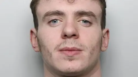 A mugshot of a 20-year-old man. He has dark brown hair which is short on the sides and curly on the top, bushy eyebrows and minimal facial hair. He appears to be smirking slightly. He is wearing a grey sweatshirt and is standing in front of a pale, plain background.