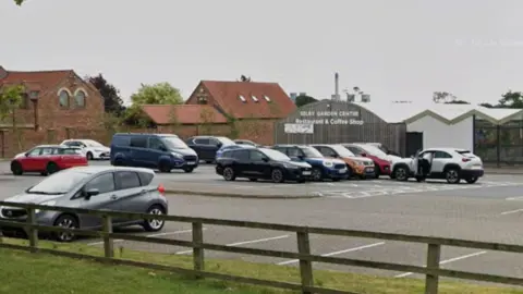 Google The exterior of a building which has a sign in white saying 'Selby Garden Centre restaurant and coffee shop'. There is a car park surrounding it with several parked cars.