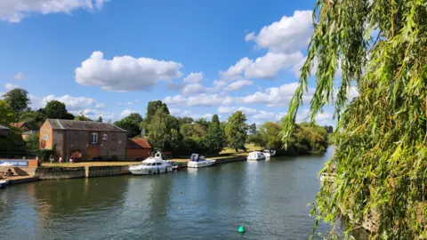 violetbloom A river with three white boats moored on the far bank. There is a brick building and the river is flanked by green trees. Overheard the sky is blue with white clouds.