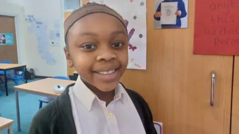 Apryl, a girl aged nine, is looking at the camera. Behind her on the left are three tables and a door and a cupboard with pictures on the doors is on the right.