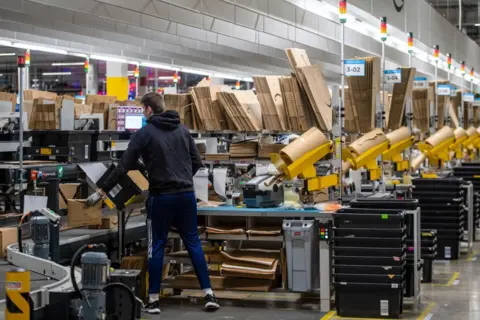 Getty Images A worker at a warehouse packages items as they pass him on a conveyor belt.