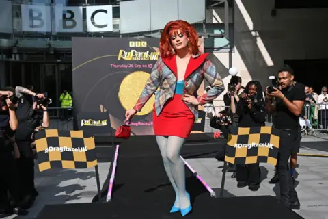 Getty Images Chanel O'Connor, a drag Queen competing on this year's Ru Paul's Drag Race UK. She is wearing a red wig and lots of makeup. She has in gold earring and is wearing a tartan blazer. She is posing at the end of a catwalk at the BBC's broadcasting house in London.