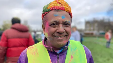 BBC Bhardwaj smiles. He is wearing a high-vis vest, a purple fleece and a brightly-coloured head covering. He has yellow, pink and blue paint smeared on his face and clothes. 