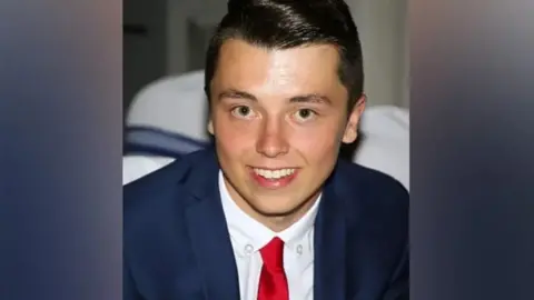 Family photo Connor Brown. He is smiling and has short black hair. He wears a blue suit, white shirt and red tie