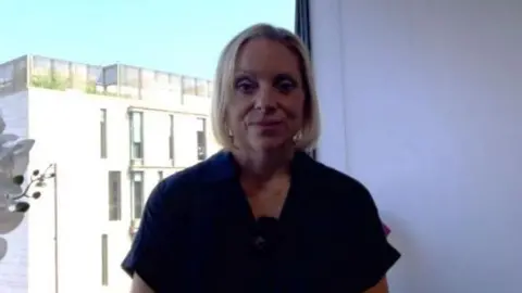 A woman with short blonde hair infront of a window showing Stafford town centre in the background.