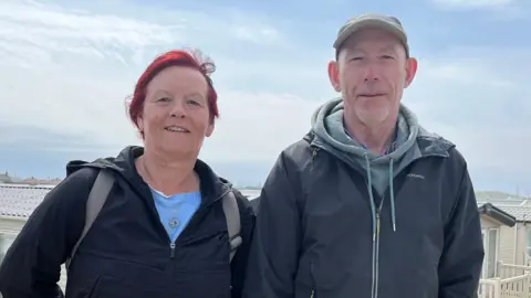 Carol and Wayne Henshaw standing outside Redcar Beach Caravan Park. They are both wearing warm jumpers and coats. Carol has short, red hair. Her husband wears a grey cap.