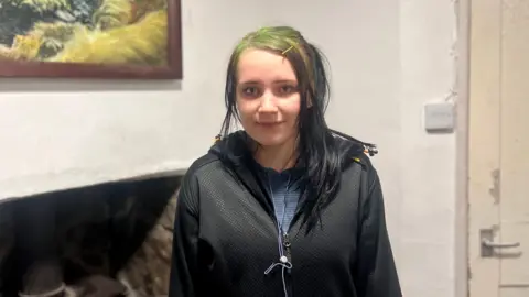 Britta smiling at the camera. She has green and black hair and is wearing a black hooded jumper. There is a painting hanging on the wall to the left.