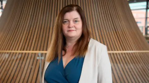 Sioned Williams, who has brown, straight hair, looks directly into the camera wearing a cream jacket and blue wrap dress.