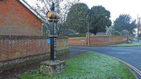 Adrian S Pye/Geograph A village sign has a cobble and brick square base with a dark wood beam with the name "Old Catton" spelt vertically in gold lettering, with a model of a black cat sat on a brown barrel at its top. The sign is shown on a grass verge next to a road, with the walls of a housing estate and trees shown behind it.