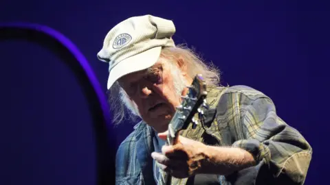 PA Media Neil Young performs in a plaid jacket and cap.