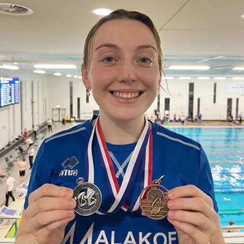 Katie Bain Katie is in the indoors swimming pull, the pool itself id behind her, she is wearing navy blue t-shirt and is holding silver medal in one hand and bronze in the other