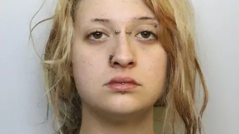 A mugshot of a young woman with blonde hair. It tied back in a messy ponytail. She has her lip, eyebrow, nose and the bridge of her nose pierced with metal bars and has a neutral expression.