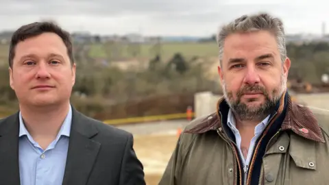 Two men, one in a black blazer and a blue shirt and the other in a Barbour-style farmer's coat, standing in front of the bypass under construction.