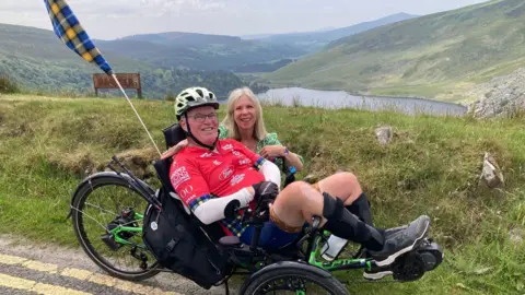 Supplied Nick Apperley sitting on a trike on the side of a road but at the top of a hill. He is wearing a red cycling top, helmet, and blue shorts and shoes. A woman is crouching down behind him. She is wearing a green and white dress. They are both smiling and looking into the camera.