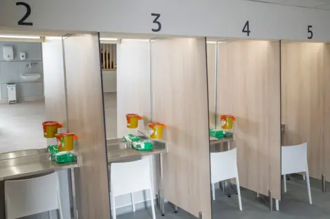 PA Media Numbered booths with a white chair in each booth. Each one faces a mirrored wall. Each booth has a green pack and a yellow sharps bin. It is brightly lit.
