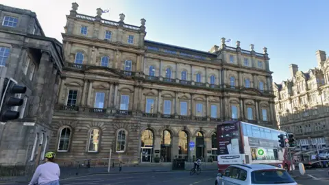 Google A large Victorian building at the east end of Princes Street in Edinburgh. There are buses and cars in front of it and the sky is blue.