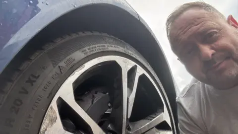Jason Conyers A man with short fair hair and a grey T-shirt crouches down by the wheel of his blue Audi. Although only part of the tyre is visible, the alloy wheel is scuffed. 
