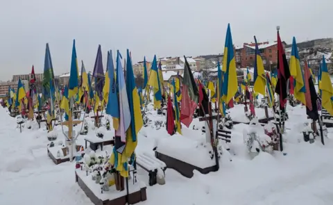 Supplied A snowy graveyard with Ukraine flags attached to each grave. There are buildings in the background.