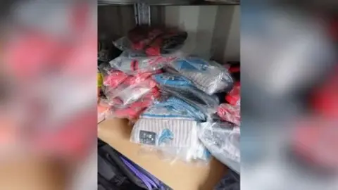 Dozens of woolly hats with NFL branding sit on a table, still in plastic packaging. Some are grey and blue, while others are red. 