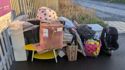 Enfys Foundation Piles of bags and boxes of donations outside the gate of the charity store including a baby car seat and yellow coffee table. 