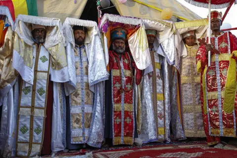 Anadolu via Getty Images A group of priests in traditional clothes hold the top of their colourful robes up above their heads.